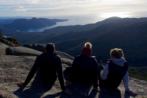 Three people sitting on a mountain looking away from the camera towards the coast